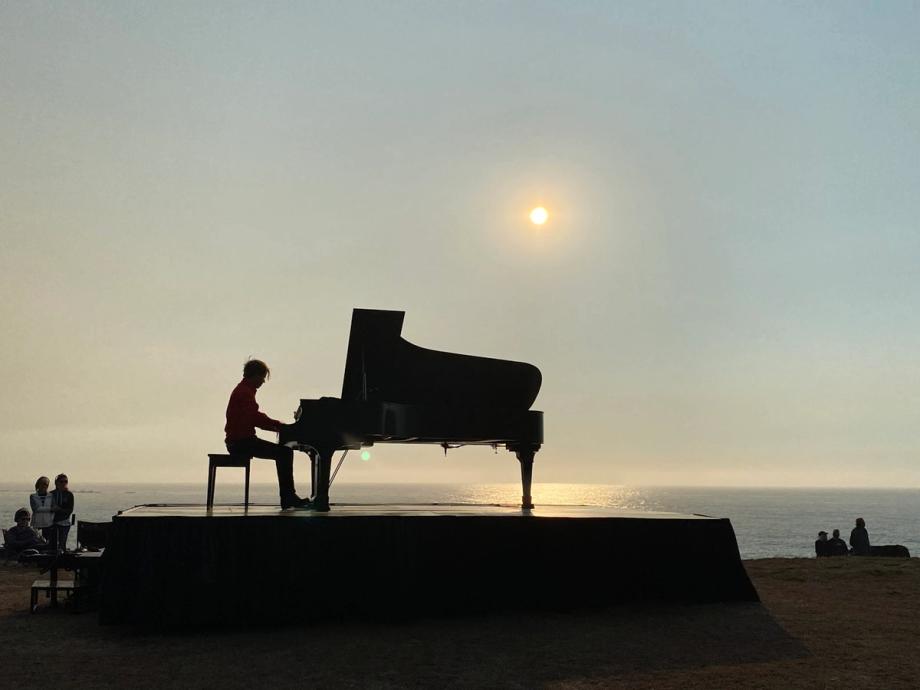 A pianist performs on a grand piano on an outdoor stage overlooking the Pacific Ocean at sunset during an In a Landscape classical music concert at Shore Acres State Park in Oregon, with small groups of audience members visible on either side.