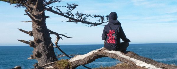 A person sits quietly on a weathered coastal tree branch overlooking the Pacific Ocean on Oregon's Adventure Coast, wearing a hoodie that reads 'Adventure.'