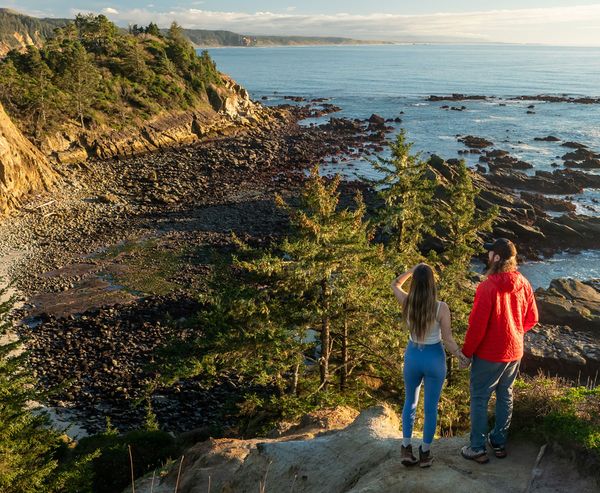 A couple stands on a cliffside overlook watching the rocky coastline and ocean at Cape Arago State Park.