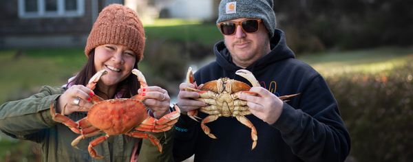 A smiling woman and man each hold up a whole cooked Dungeness crab outdoors.