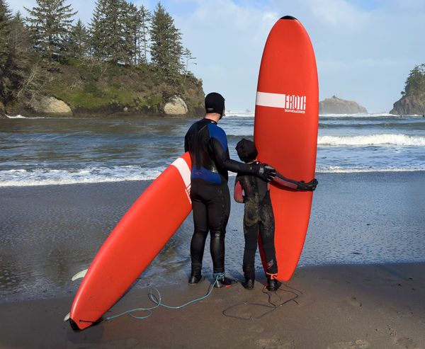 Adult and child in wetsuits holding red surfboards, looking out at the ocean and sea stacks at Sunset Bay.