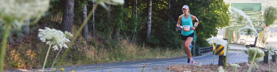 A woman runner with a race bib passes a green bridge along a tree-lined road during the Circle the Bay Relay.