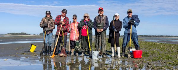Seven clammers pose together on a tidal flat near Charleston, Oregon, holding shovels and clam guns with buckets and a net at their feet after a successful dig.