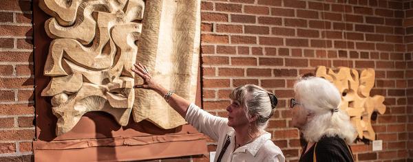 Two women examine an abstract wood relief sculpture mounted on a brick wall at the Coos Art Museum, with one reaching out to touch the carved surface.