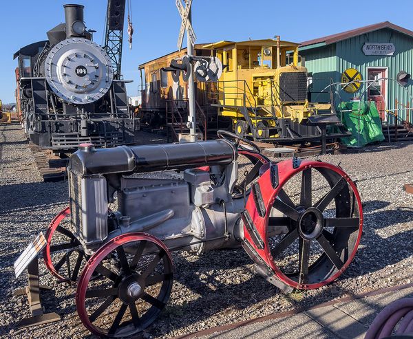 A vintage tractor with red wheels on display at the Oregon Coast Historical Railway Museum, with steam locomotive No. 104 and a yellow rail crane visible behind it.