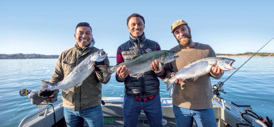 Three smiling anglers hold up freshly caught Chinook salmon aboard a charter fishing boat near Coos Bay, Oregon, under a clear blue sky.