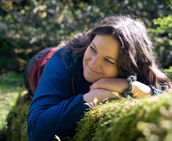 A smiling woman resting her chin on her hands on a moss-covered log in a sunlit forest.