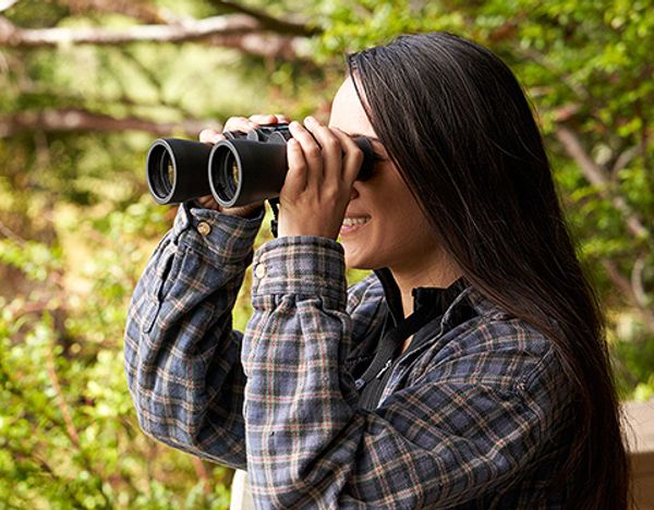A woman smiling while looking through binoculars in a forested area.