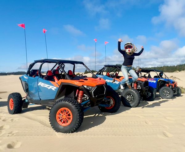 Three orange and blue RZR side-by-sides lined up on a dune, with a helmeted rider jumping joyfully beside them.