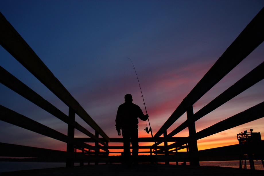 Silhouette of a person fishing from a pier at sunset, with a colorful orange and pink sky over the water.