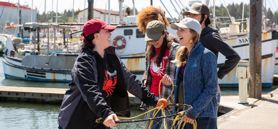 A group of women laughing together on the dock at Charleston Marina, holding a crab ring net beside moored fishing boats.