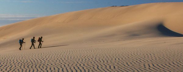 Three hikers with backpacks and trekking poles cross a rippled sand dune at Oregon Dunes National Recreation Area, casting long shadows in late-day light.