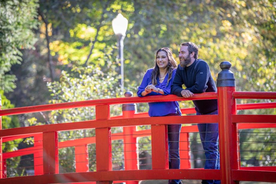 A smiling couple leans against the railing of a red arched footbridge in a garden, surrounded by trees in Coos Bay’s Mingus Park.