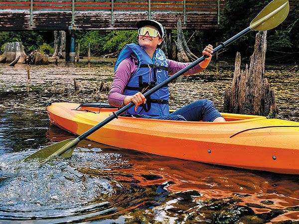 A woman wearing a life jacket and sunglasses smiles while paddling an orange kayak through a calm, forested waterway beneath a wooden bridge