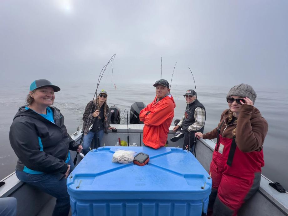 A group of five anglers on a boat with fishing rods deployed, surrounded by fog and calm water