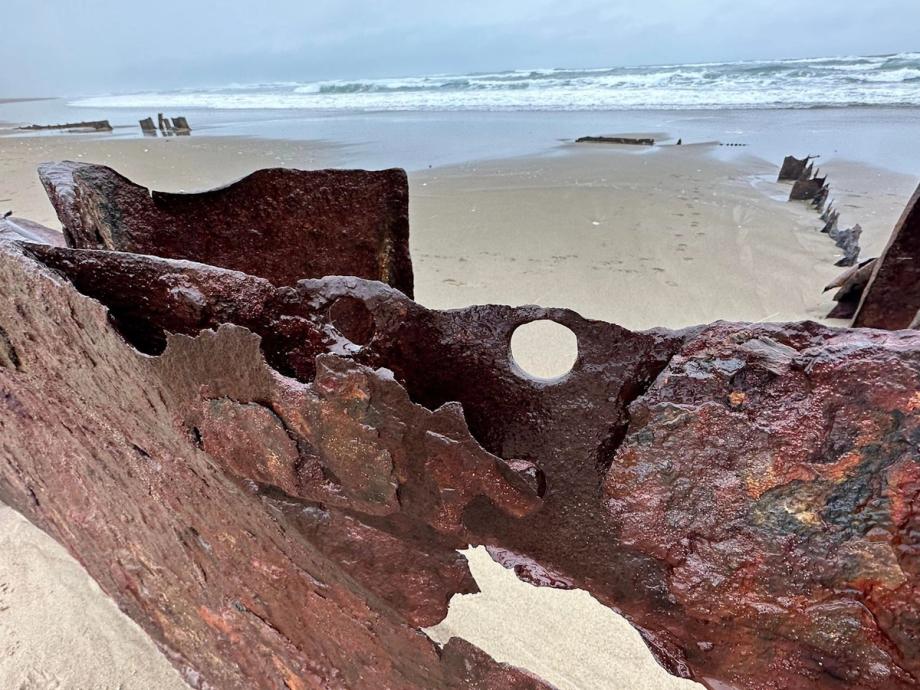 Close-up of large rusted, weathered metal shipwreck wreckage from the 1929 wreck of the Sujameco, jutting from Horsfall Beach, with more scattered wreckage fragments and ocean waves in the background.