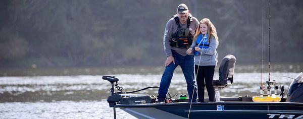 An adult and a young girl stand together on a bass fishing boat on a calm inland lake, smiling as she reels in a fish on a misty morning.