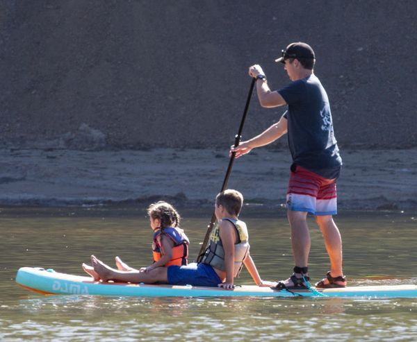 Teenager paddleboarding while two children ride along on the board near a bank in Sunset Bay.