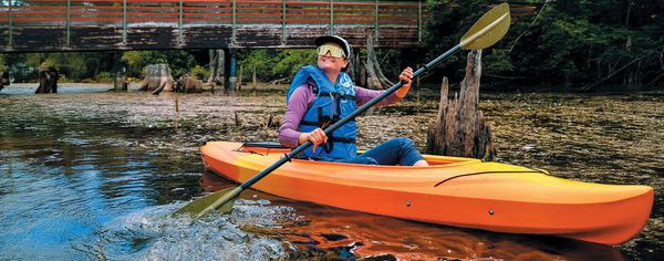 oman smiling while paddling an orange kayak under a wooden bridge through calm water.