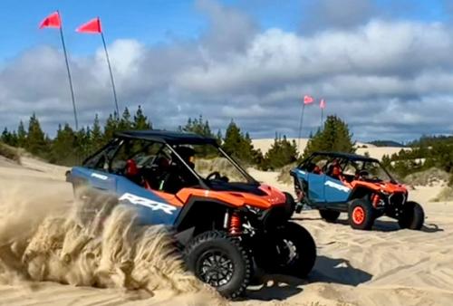 Two off-road buggies with tall safety flags kick up sand while driving through a dune area near a tree line.