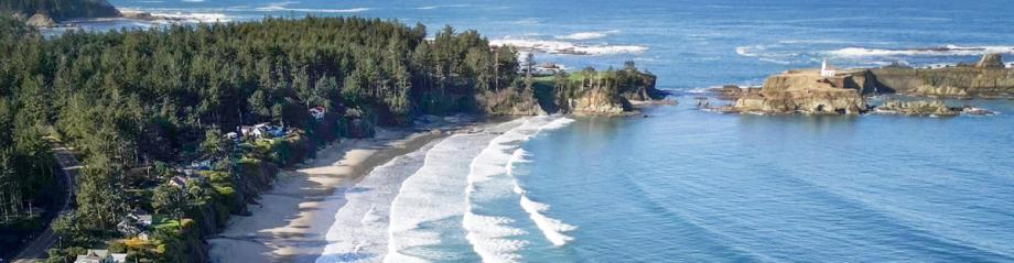 erial view of Lighthouse Beach with a curved sandy shoreline, rolling waves, forested cliffs, and a lighthouse on a rocky point in the distance.