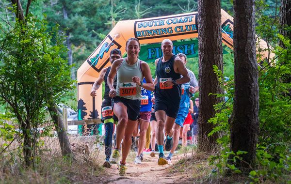 Runners run through a South Coast Running Club starting arch on a wooded trail.