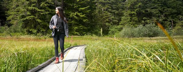 A woman with binoculars walks along a wooden boardwalk through a grassy marsh at the Estuary Study Trails.