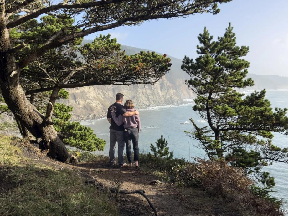 A couple stands with their arms around each other on a coastal overlook, framed by windswept pine trees, gazing at dramatic ocean cliffs and blue water along the Cape Arago Highway in Oregon.