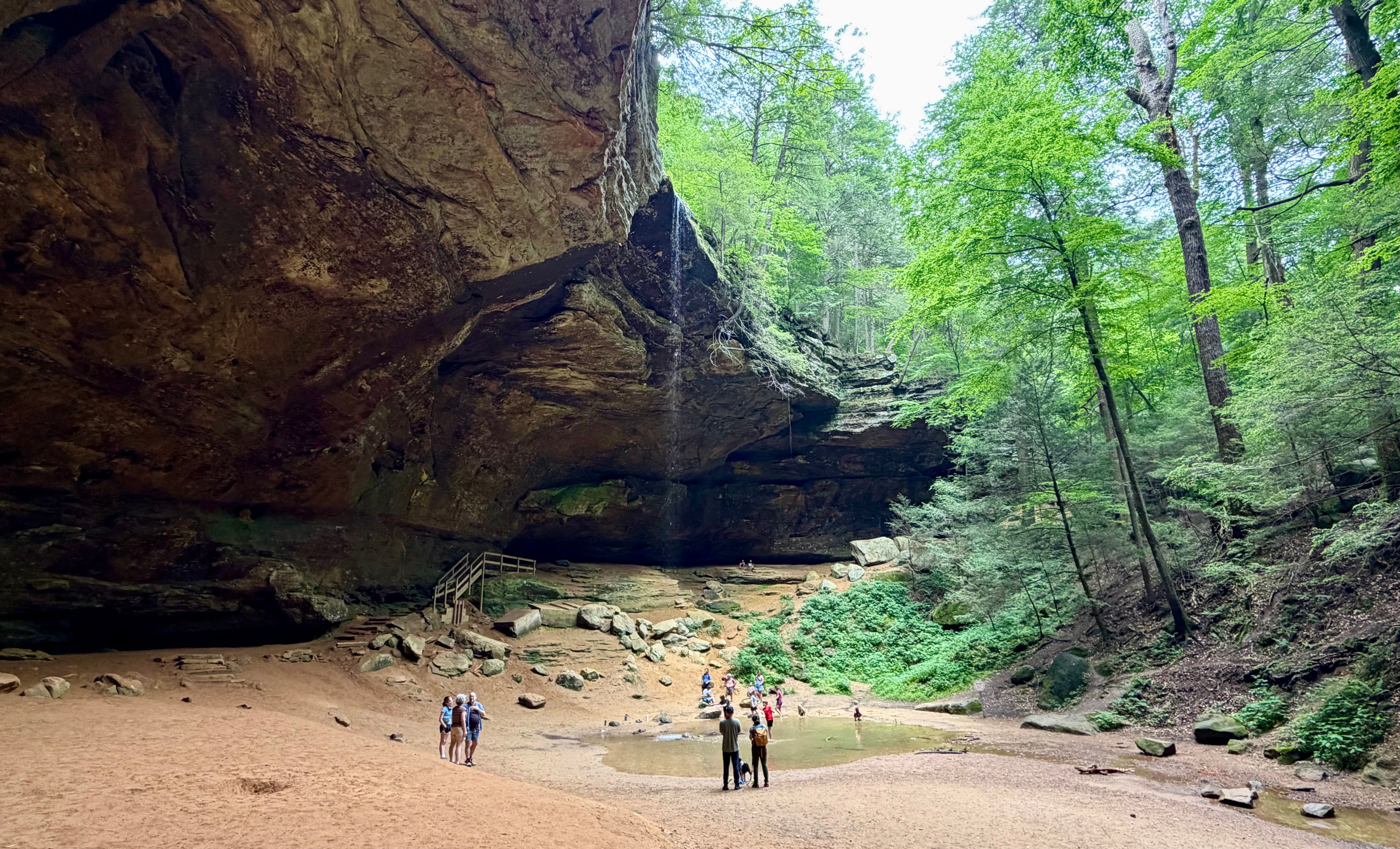 Visitors gather beneath the enormous sandstone recess of Ash Cave as a narrow waterfall drops into a shallow pool.