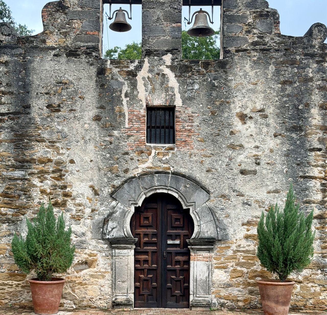 Dark wooden double doors beneath a rounded stone arch, with two bells hanging above in Mission Espada’s weathered façade.