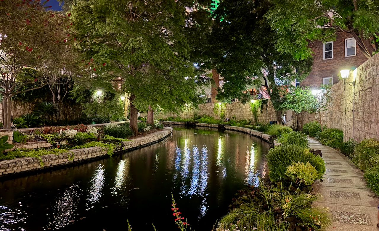 A calm bend in the San Antonio River Walk at night, bordered by stone paths, landscaping, trees, and reflected lights.
