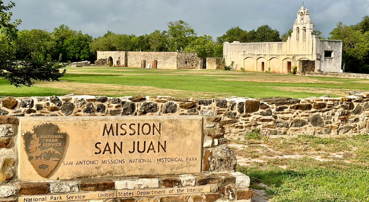 Mission San Juan entrance sign in front of a broad grassy courtyard, historic stone buildings, and the white mission church beneath a dark sky.
