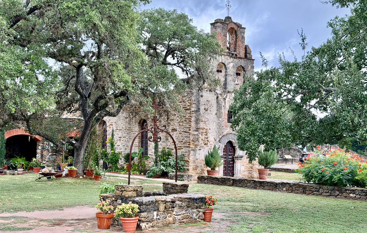Mission Espada church framed by large live oak trees, flowering plants, and a stone well under a cloudy sky.