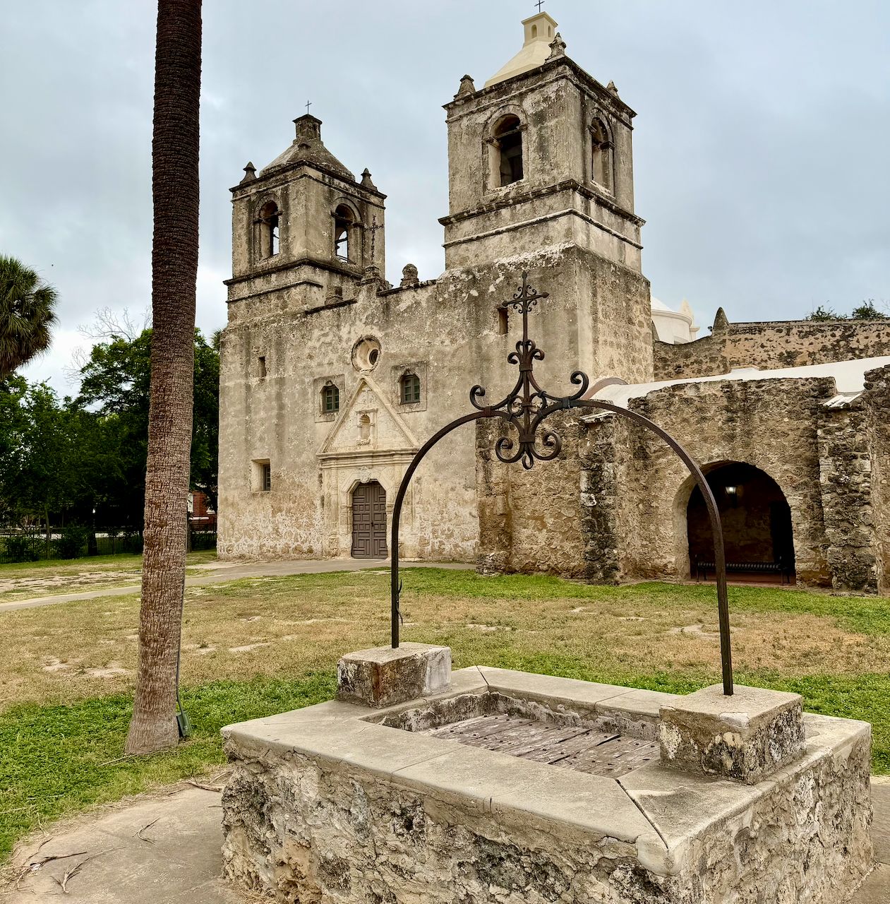 Mission Concepción church with twin stone bell towers, a palm tree, and a historic stone well beneath a cloudy sky.