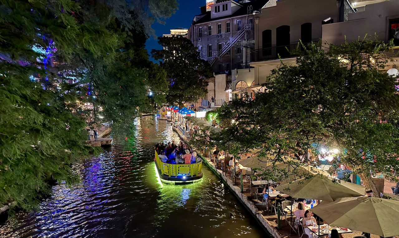 A green tour boat glowing with purple lights passes outdoor restaurant tables on the San Antonio River Walk at night.