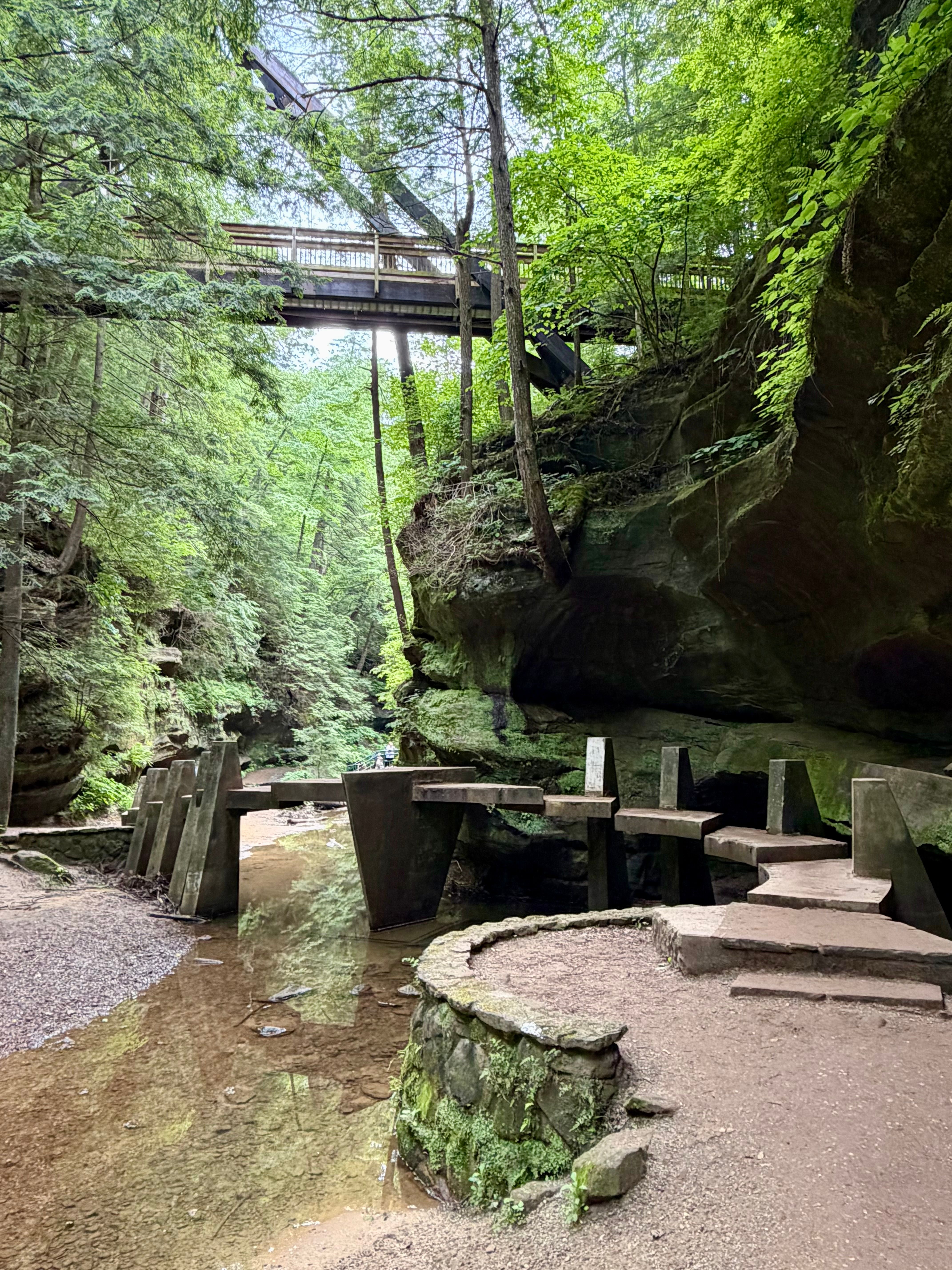 A unique concrete footbridge crosses the shallow stream in the shaded gorge along the Old Man’s Cave trail, with towering sandstone cliffs and an elevated walkway visible above.