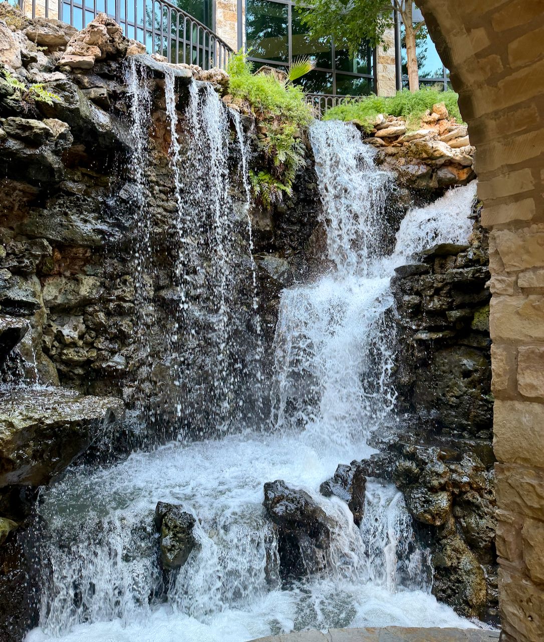 An artificial waterfall cascades over dark rocks beside a stone arch along the San Antonio River Walk.