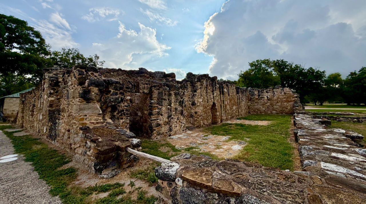 Weathered stone ruins at Mission San Juan with grass inside the former rooms and sunlight breaking through large storm clouds.