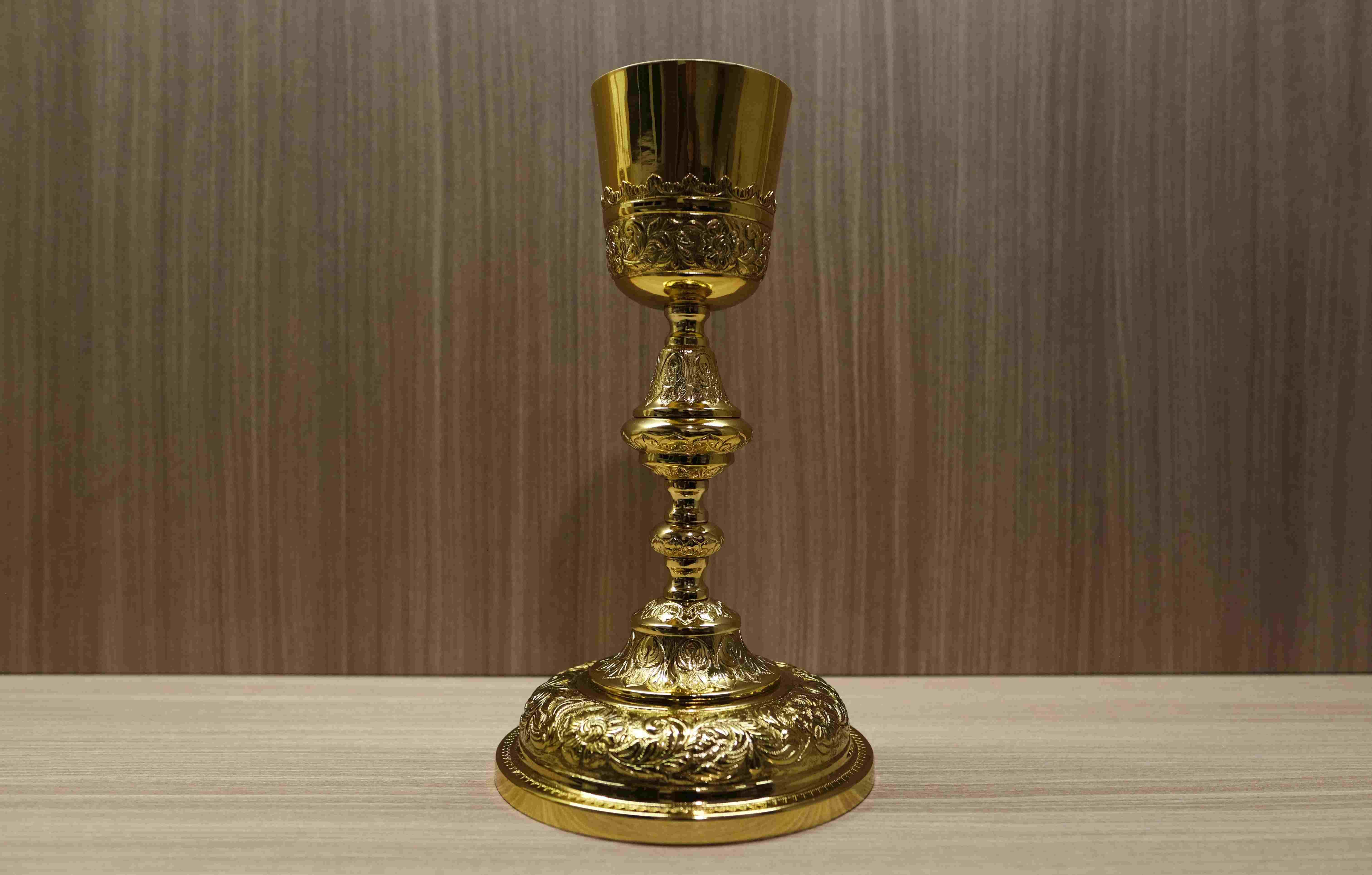A highly detailed, ornate gold chalice stands on a wooden surface against a backdrop of textured brown and tan wallpaper. The chalice has intricate, decorative carvings on its base, knop (the node in the middle of the stem), and the lower part of the cup. The upper portion of the cup is smooth and reflective. The lighting highlights the gilded surfaces and the detailed craftsmanship of the sacred vessel.