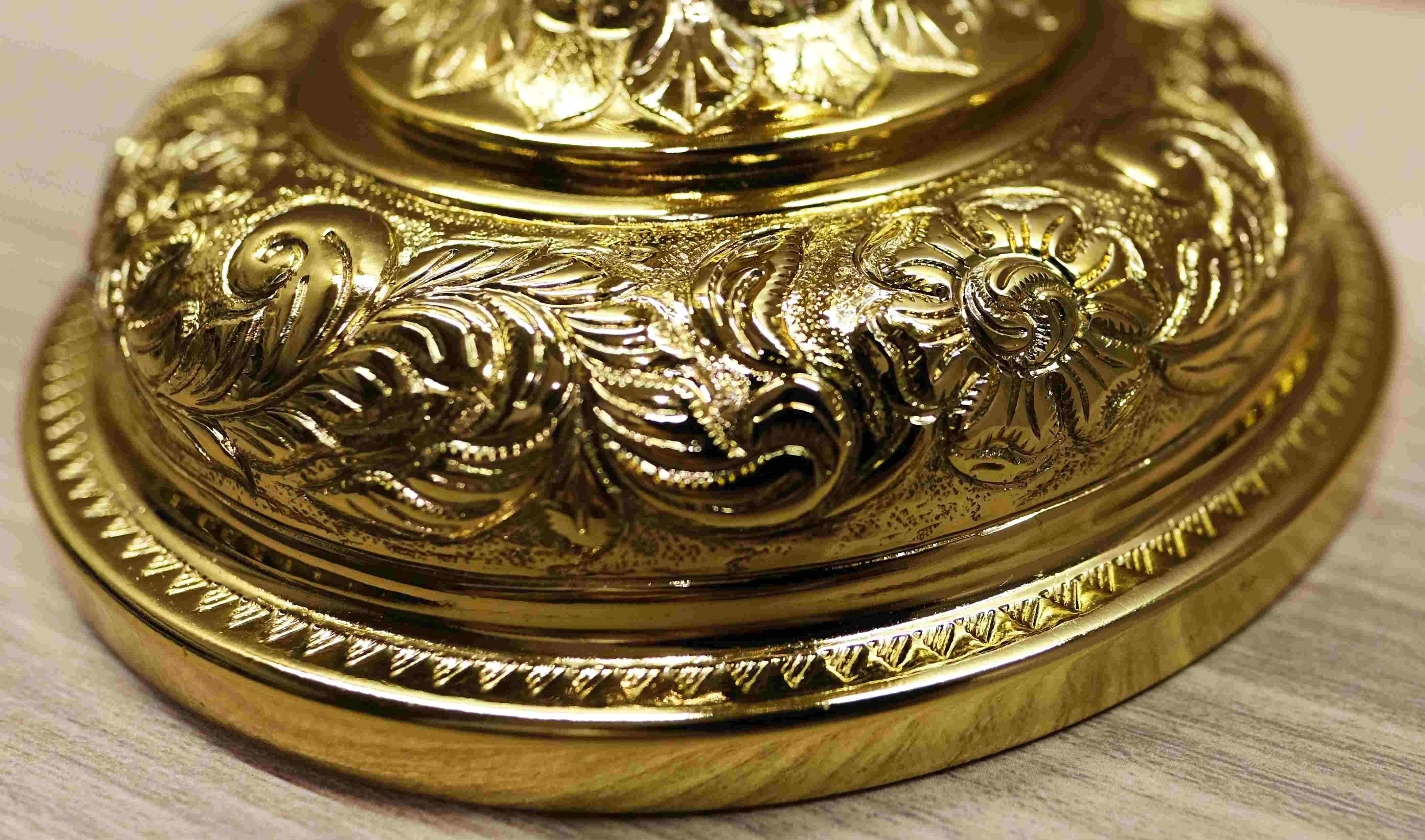 A close-up shot of the ornate, gilded base of a chalice. The circular base is highly detailed with intricate floral and scrollwork carvings. The light reflects off the polished gold surface, emphasizing the fine craftsmanship and textures. The chalice sits on a wooden surface, with a blurred background.