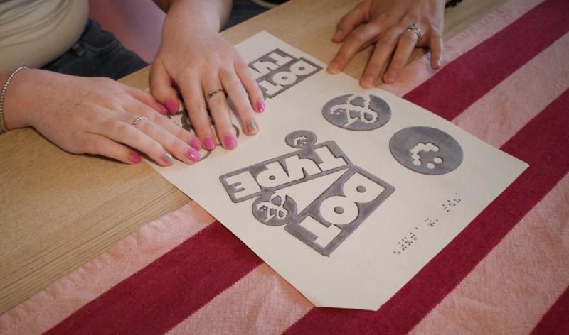 Two sets of hands reading a braille printed poster laid on a desk