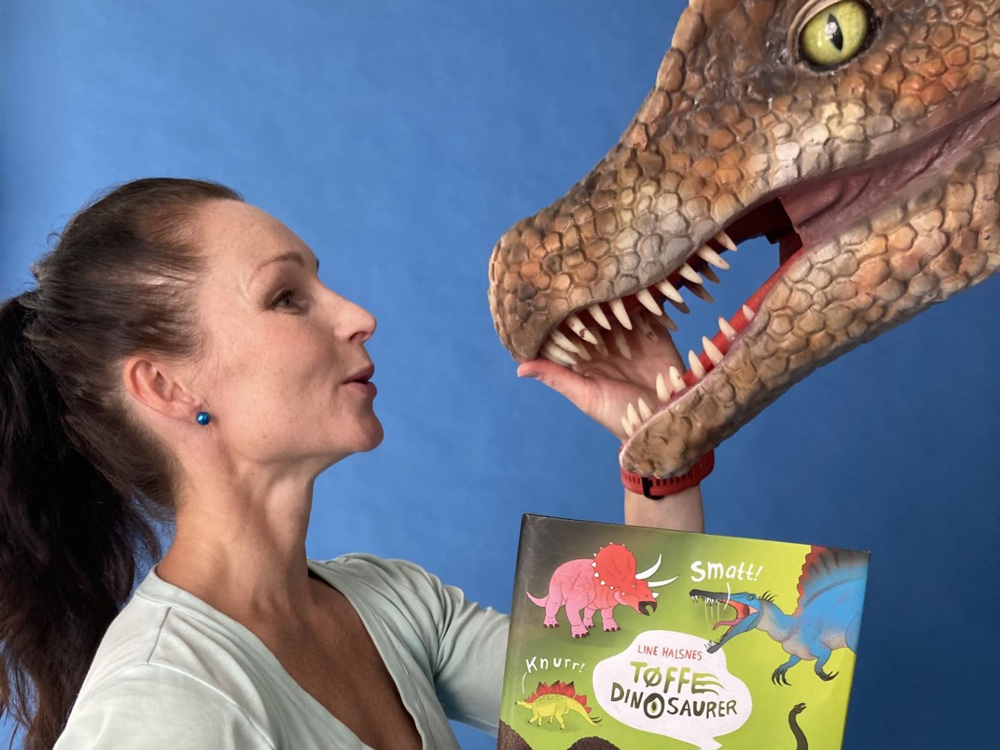 Forfatter Line Halsnes holder en bok om dinosaurer og klapper på en stor dinosaurdukke.
