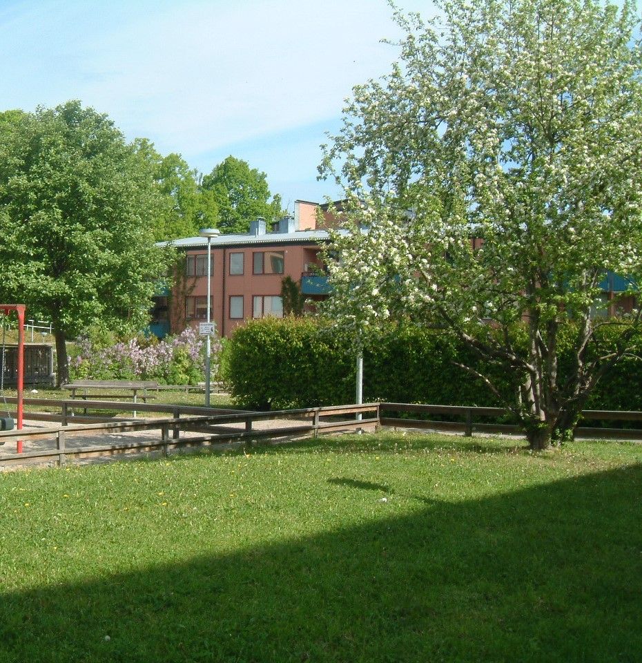 Grönområde i Ekebyhovsvägen med ett blommande träd på höger sida, bänkar och en asfalterad yta omgiven av buskar och träd. I bakgrunden syns en byggnad med röda väggar och flera fönster under en klar himmel med några lätta moln.