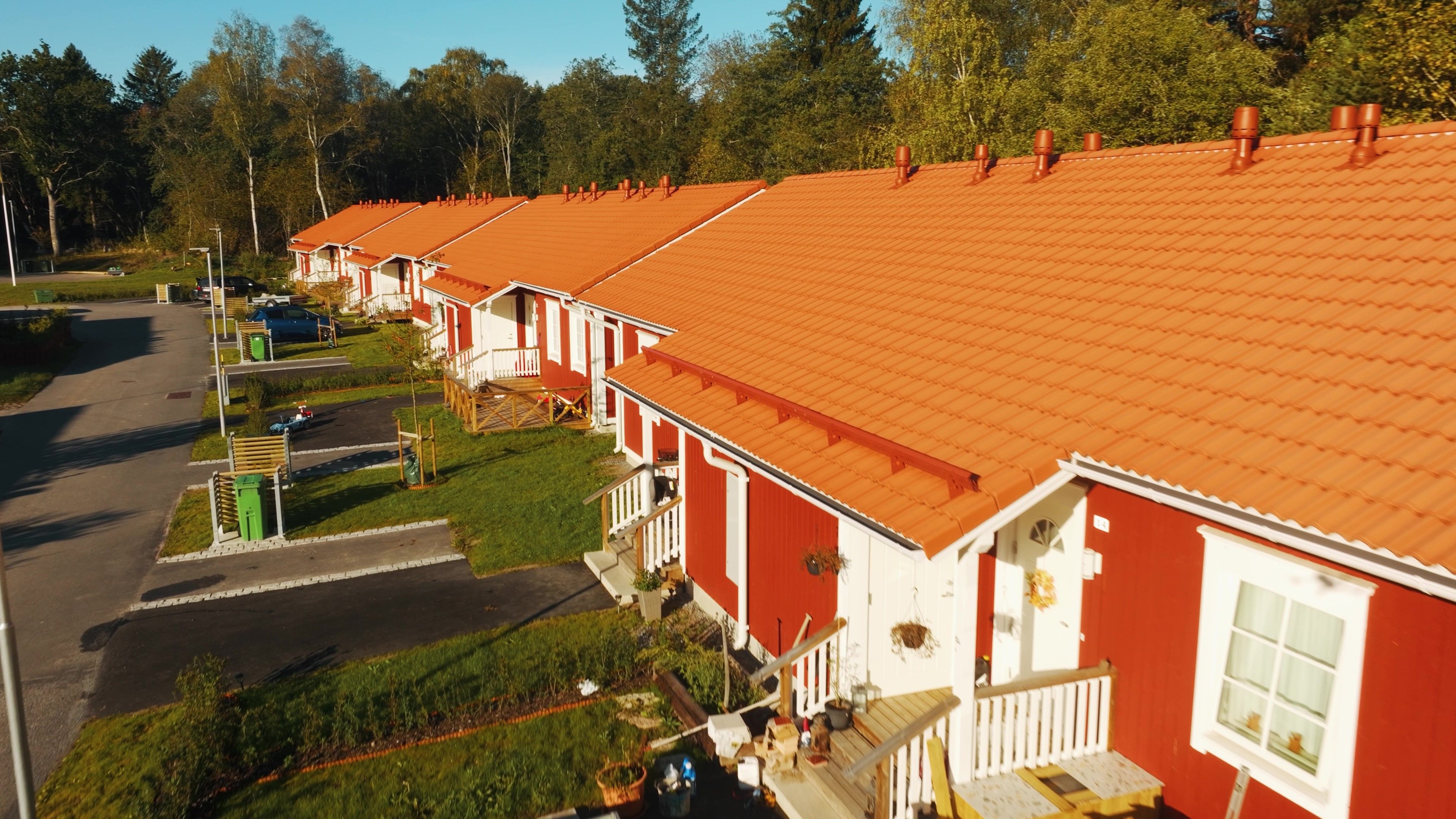 Trähus med tegeltak och två ingångar, var och en med egna trappsteg. Två gröna soptunnor står på vänster sida av huset och en på höger sida. En ljusblå vintagebil är parkerad på höger sida nära en gatlykta. Huset är omgivet av gräs, och ett liknande rött hus är delvis synligt till höger. Utomhusmöbler, inklusive stolar och bord, finns framför varje ingång.