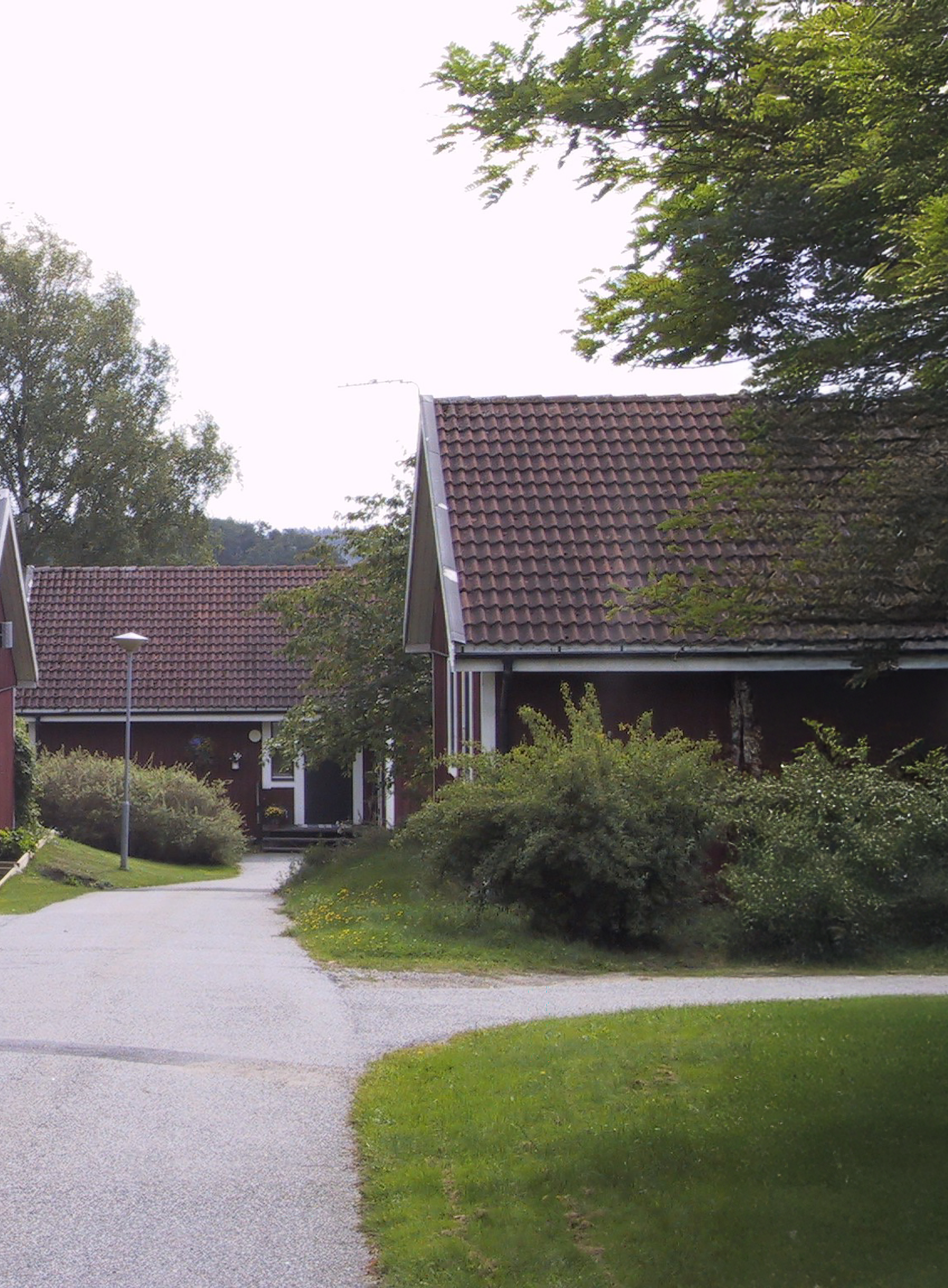 Bostadsområde med hus som har röda väggar och bruna tegeltak. En asfalterad gångväg leder genom området, omgiven av grönska inklusive buskar och träd. Himlen är mulen.