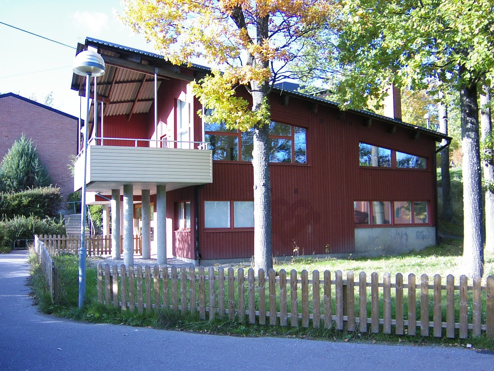 Röd träbyggnad avsedd för att vara kvarterslokal med sluttande tak och stora fönster, omgiven av träd med höstlöv. Byggnaden har en balkong på vänster sida, stödd av pelare, och en extern trappa som leder upp till den. Framför byggnaden finns ett trästaket.