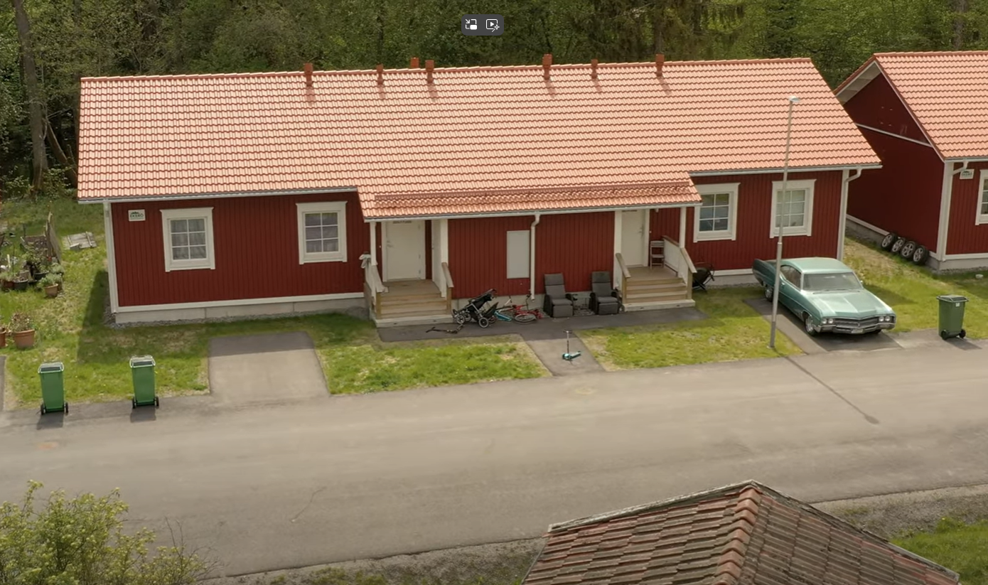 Trähus med tegeltak och två ingångar, var och en med egna trappsteg. Två gröna soptunnor står på vänster sida av huset och en på höger sida. En ljusblå vintagebil är parkerad på höger sida nära en gatlykta. Huset är omgivet av gräs, och ett liknande rött hus är delvis synligt till höger. Utomhusmöbler, inklusive stolar och bord, finns framför varje ingång.