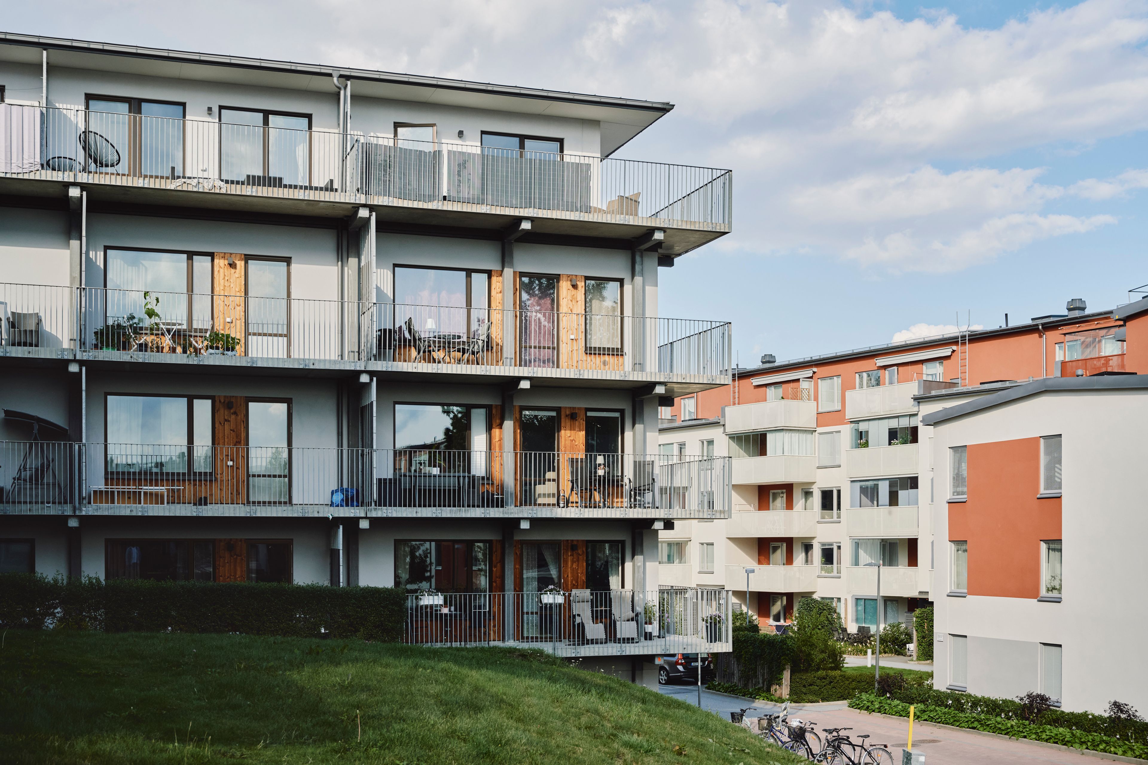 Flerbostadshus med fyra våningar, där varje våning har flera lägenheter med balkonger och metallräcken. Byggnaden har en modern design med stora fönster och trädörrar till varje lägenhet. På balkongerna finns olika föremål som växter, stolar och bord. Byggnaden är belägen på ett grönområde under en delvis molnig himmel.