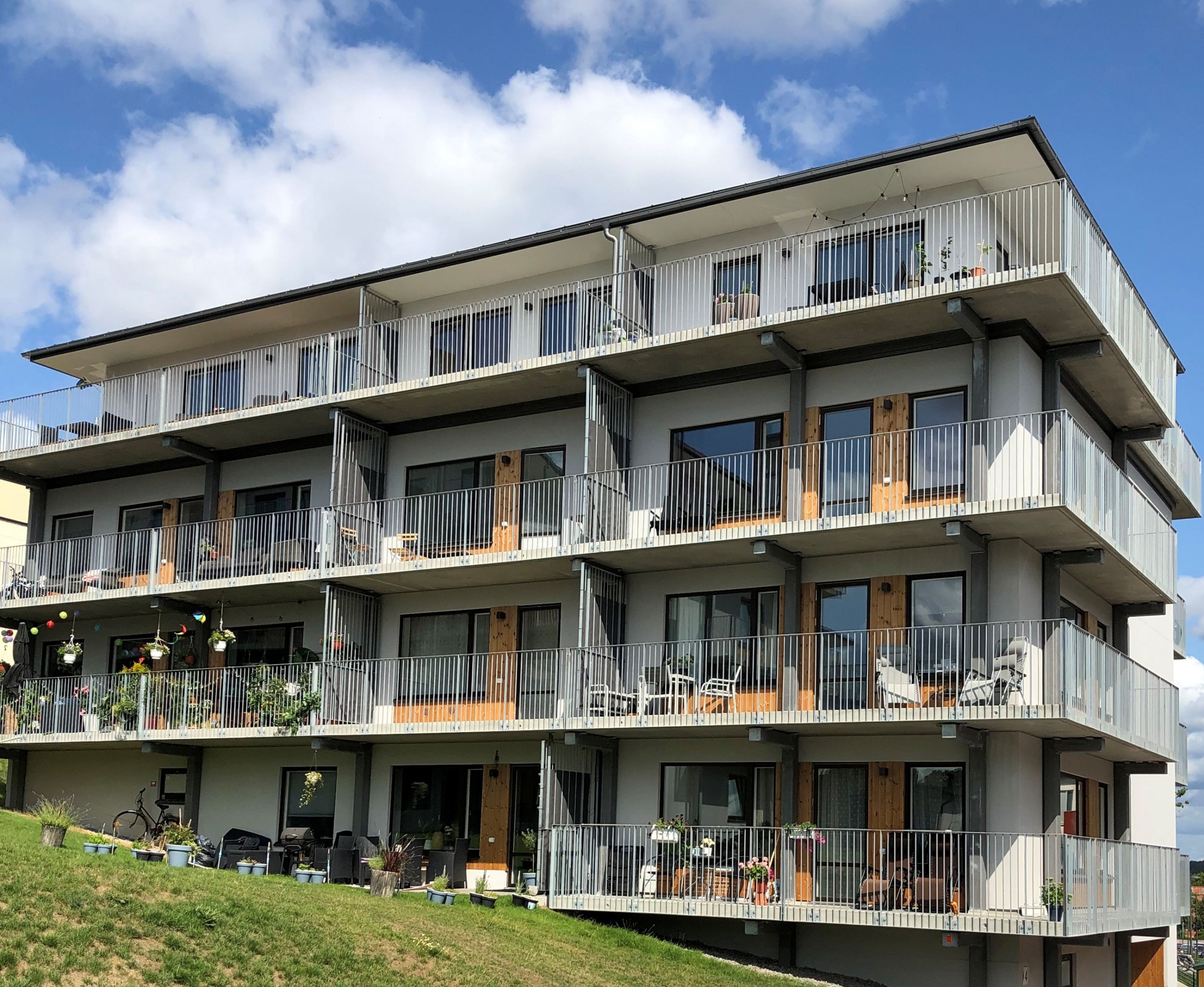 Flerbostadshus med fyra våningar, där varje våning har flera lägenheter med balkonger och metallräcken. Byggnaden har en modern design med stora fönster och trädörrar till varje lägenhet. På balkongerna finns olika föremål som växter, stolar och bord. Byggnaden är belägen på ett grönområde under en delvis molnig himmel.