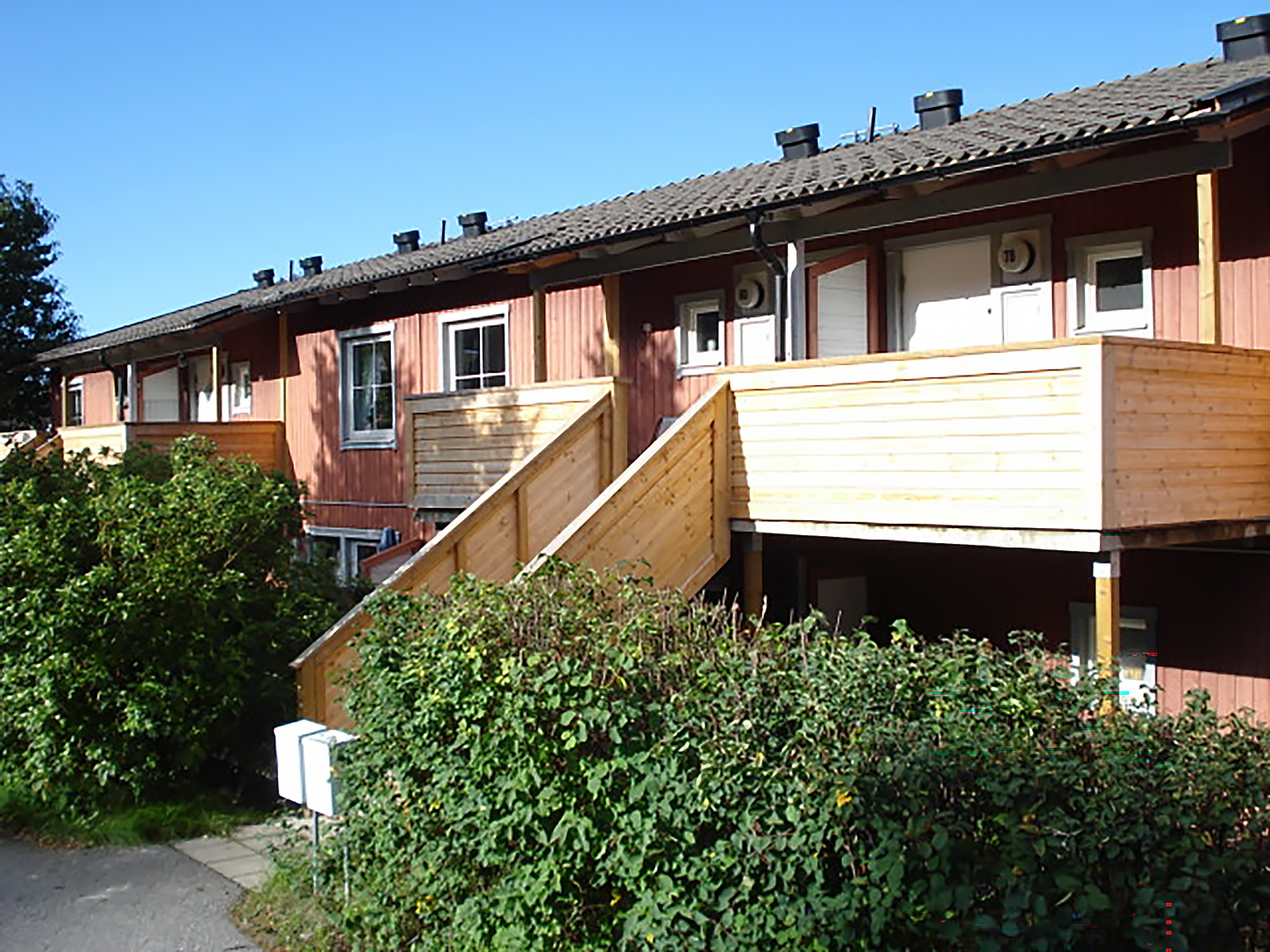 Rad av tvåvåningsradhus med träfasader. Byggnaderna har röda ytterväggar och bruna tak med flera skorstenar. Trappor i trä leder upp till ingångarna på andra våningen, och grönska, inklusive buskar, finns framför byggnaderna. Himlen är klar och blå.